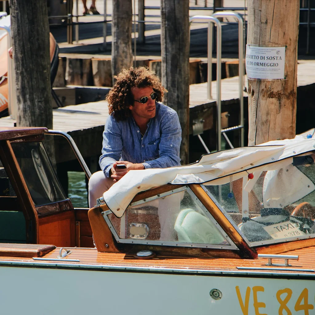 Uomo con camicia di lino azzurra e occhiali da sole vintage artigianali su barca in legno ormeggiata a Venezia, atmosfera estiva italiana.